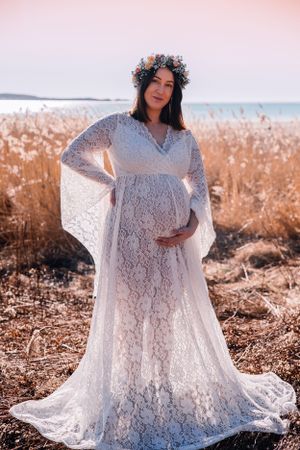 A pregnant person in a white lace maternity dress with bell sleeves and floral crown poses in a field at sunset.