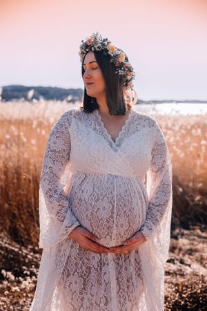 A person in a white lace maternity dress with a floral crown poses in a field at sunset during a photoshoot.
