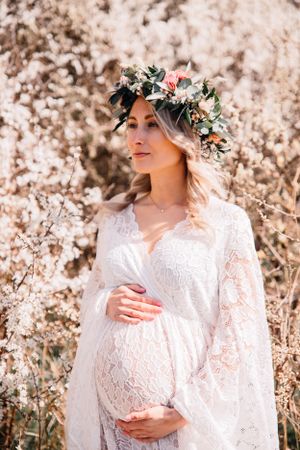 Ethereal maternity photo in white lace dress and floral crown against blooming spring background.
