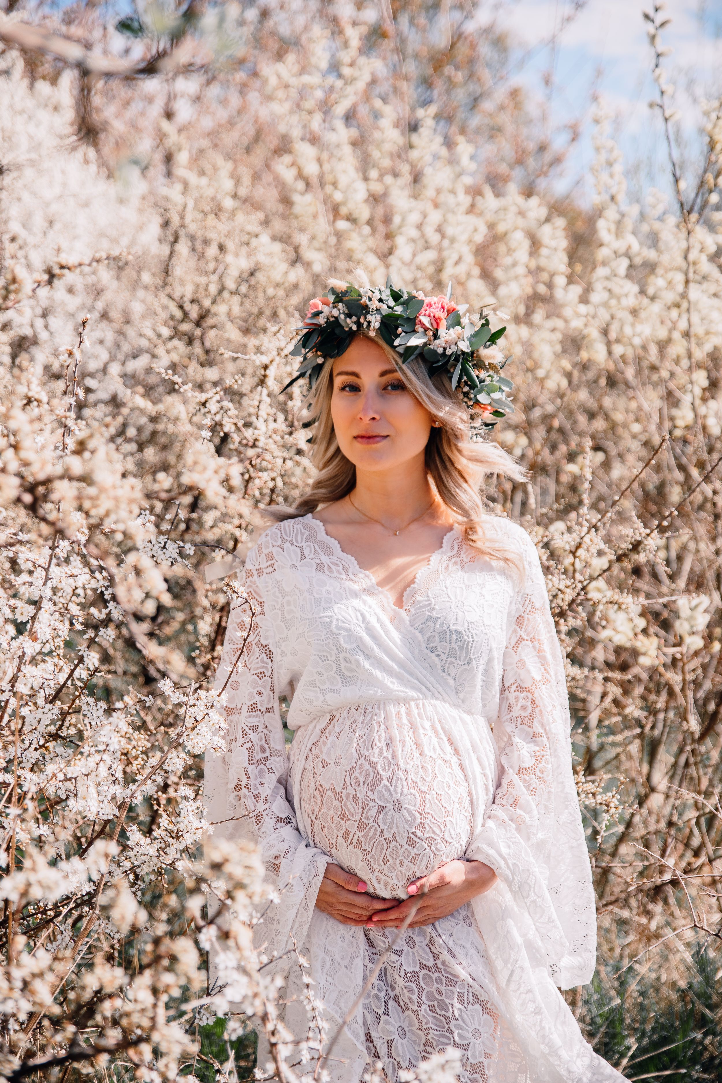 Expectant mother in delicate white lace gown wearing floral crown poses among spring blossoms.