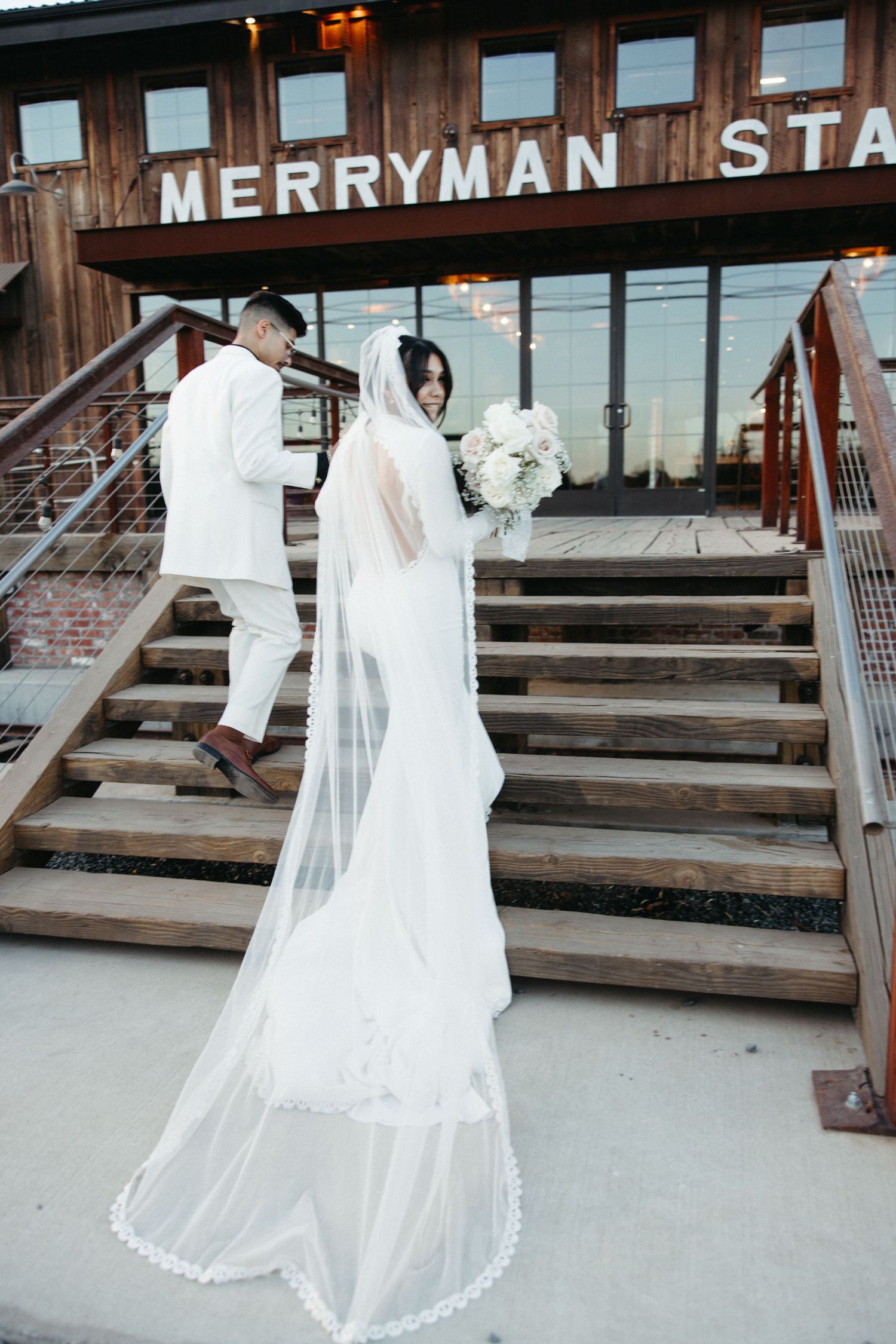 A bride in a flowing white wedding gown with long veil stands on wooden stairs outside Merryman Station.