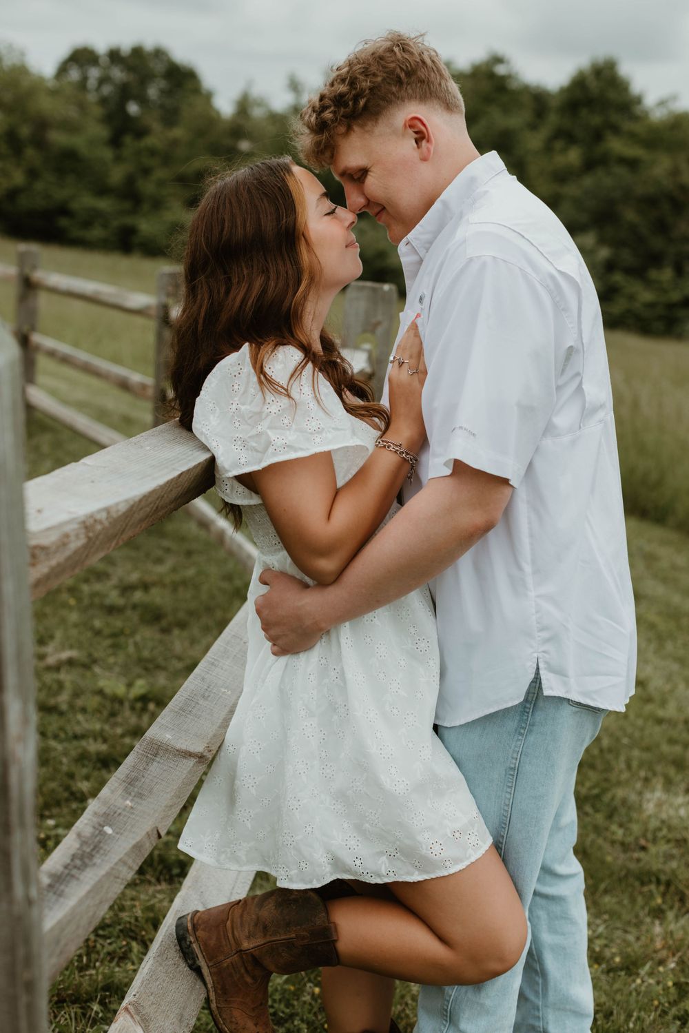 Romantic couple in white clothing embracing by wooden fence in countryside setting.