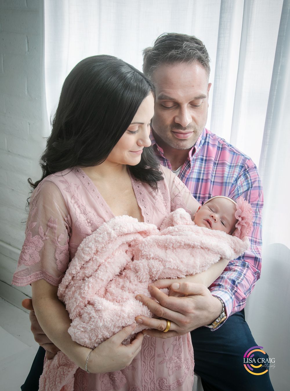 Parents share tender moments with their newborn baby in soft pink and plaid clothing near a bright window.