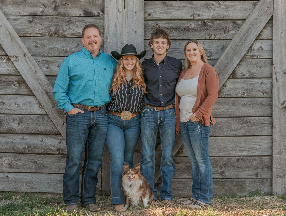 Family poses for casual outdoor portraits in front of rustic barn door wearing coordinated outfits with their Sheltie dog.