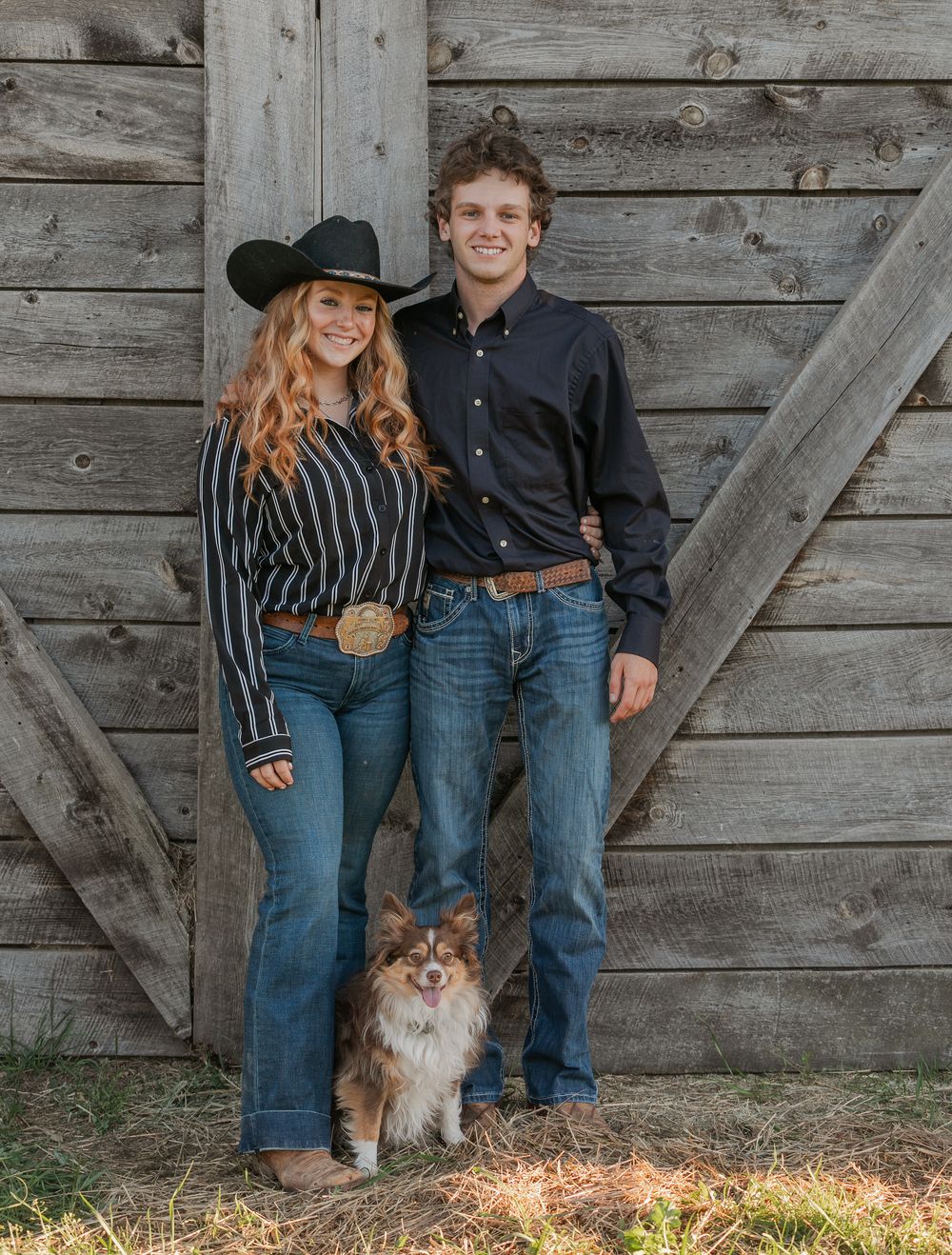 A couple and their dog pose together against a rustic wooden barn wall in matching casual denim outfits.