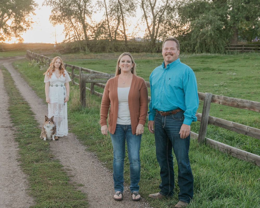 A couple and their dog walk together along a rustic country path at sunset near wooden fences and tall grass.