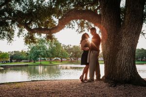 A couple embraces under a large oak tree at sunset beside a tranquil lake.