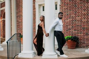 A couple poses playfully on opposite sides of a white column against a brick building exterior while holding hands.