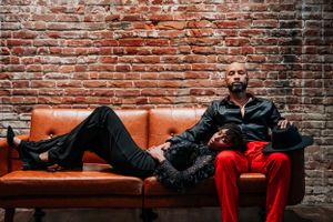 A relaxed couple lounges on a vintage leather couch against an exposed brick wall with moody lighting.
