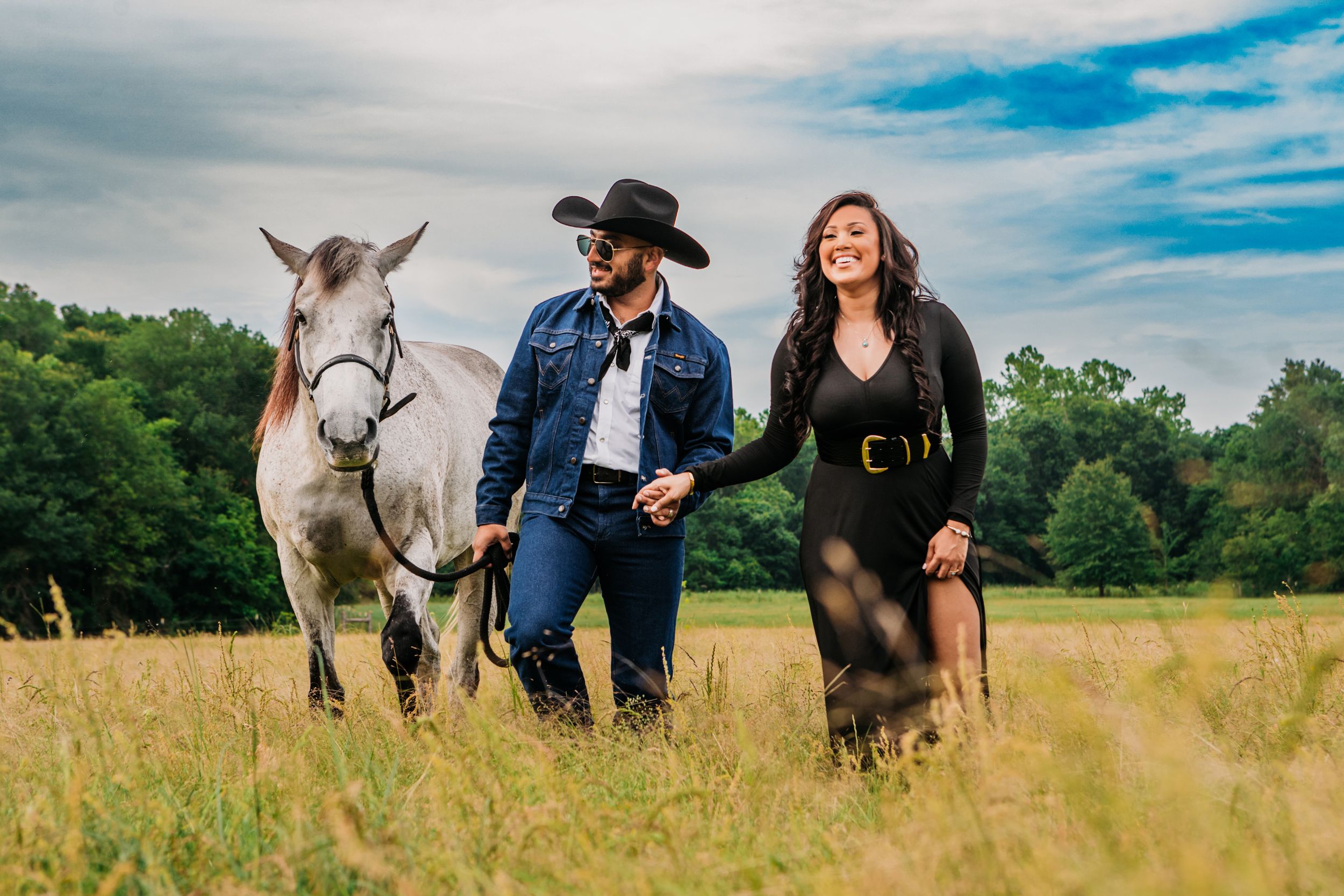 A couple stands with a white horse in a golden field on a sunny day with blue skies and green trees in background.