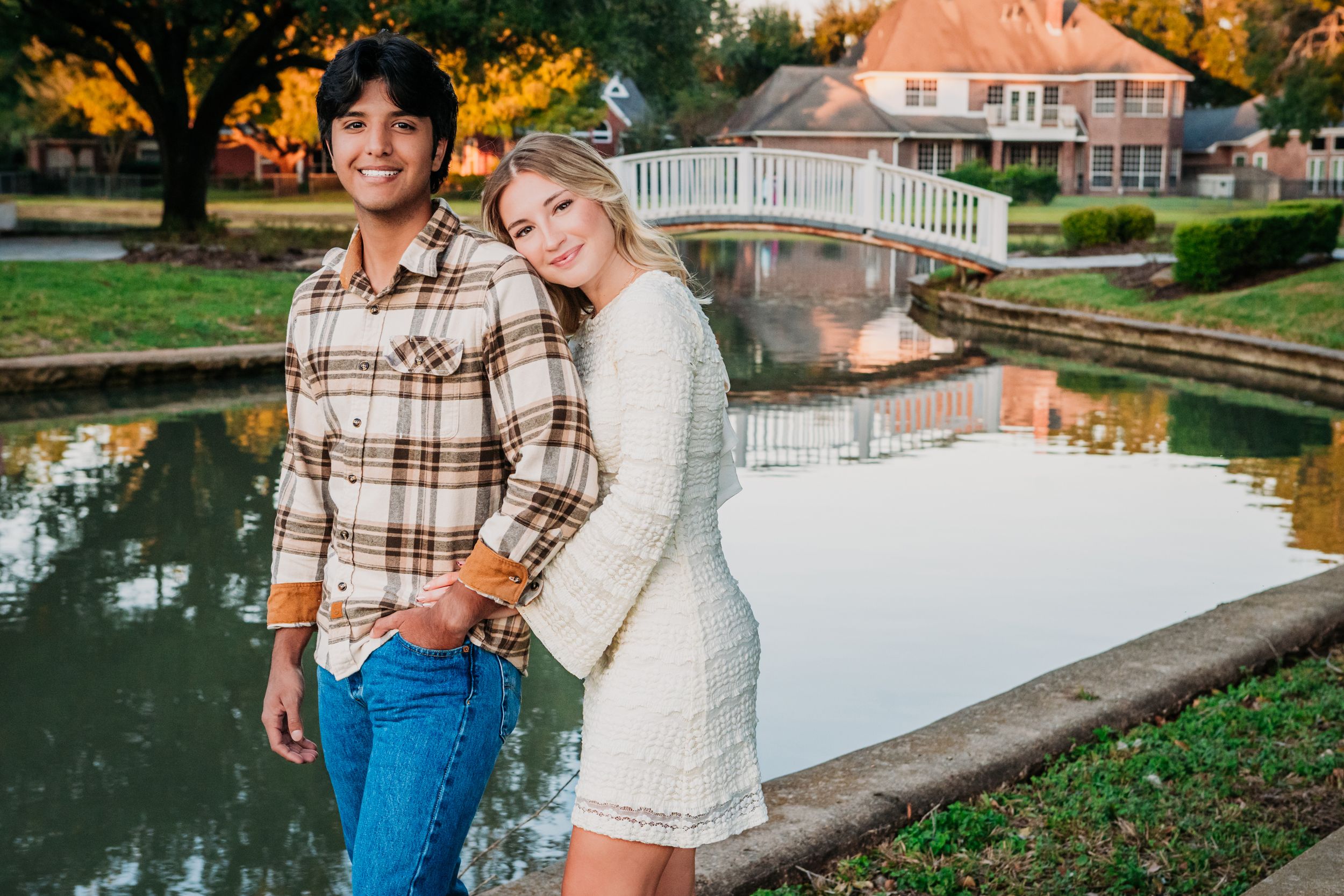 A couple poses near a scenic pond with a white bridge and autumn trees reflecting in the calm water. Engagement