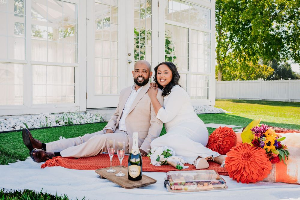 A couple lounges on orange blankets in a garden setting during a romantic outdoor photoshoot. Engagement