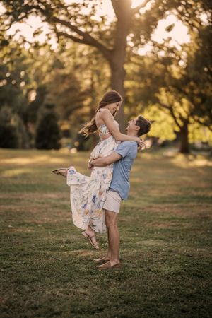 Romantic couple embraces in a sun-dappled park at golden hour as her floral dress flows in the warm evening light.