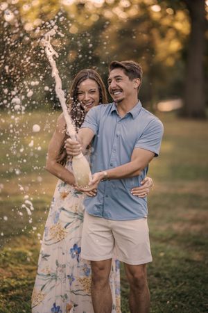 Joyful couple celebrating outdoors with champagne spray at sunset in a park setting.