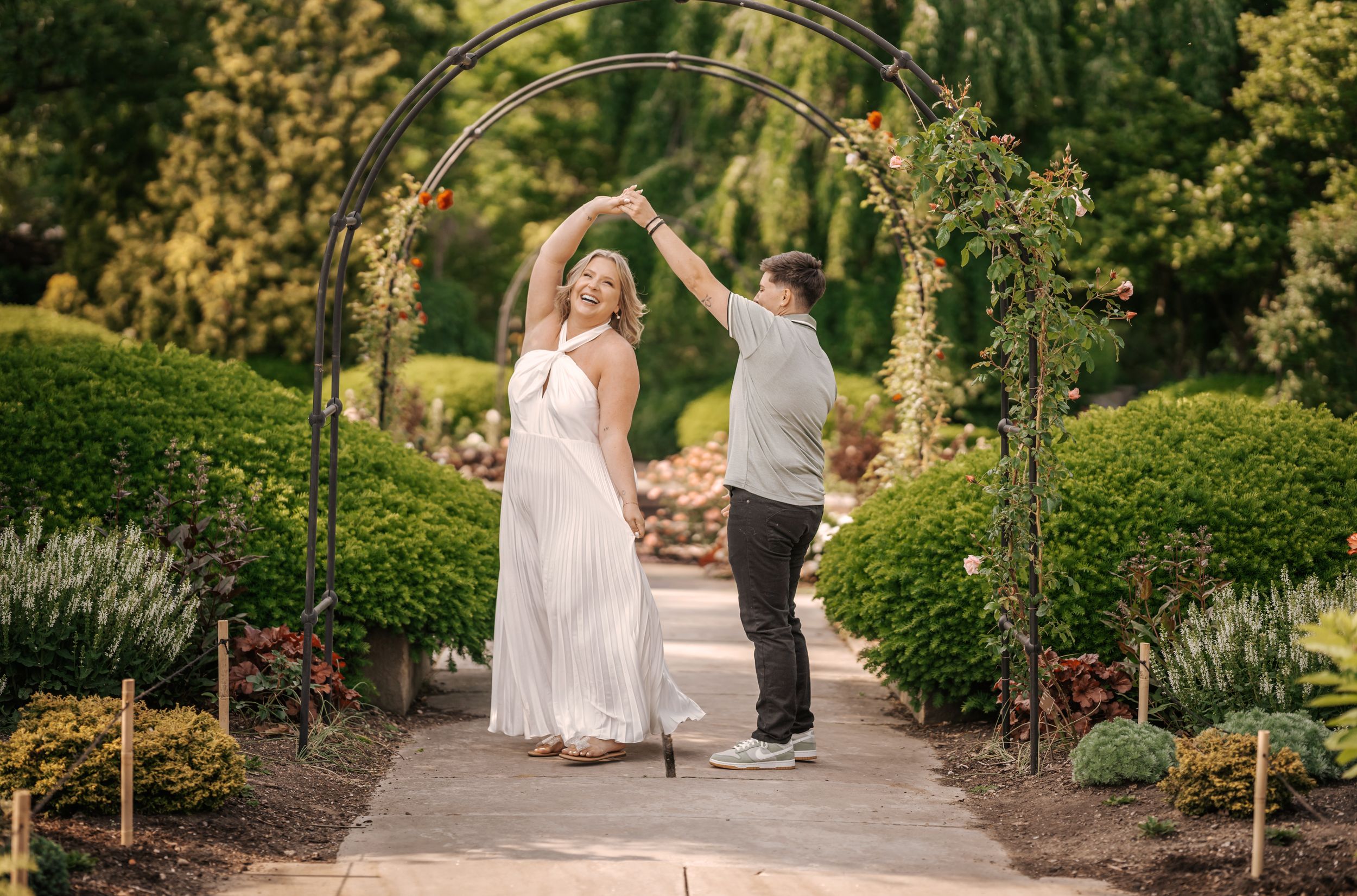 A couple dances together on a winding garden path surrounded by lush greenery and blooming flowers.