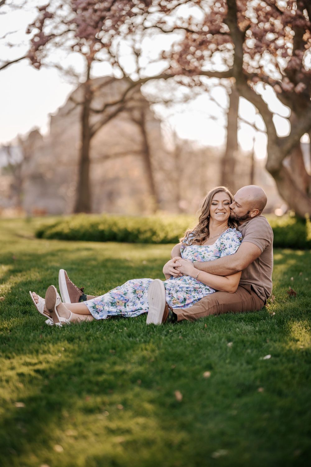 A couple relaxes together on a grassy hill during golden hour with blooming cherry blossom trees in the background.