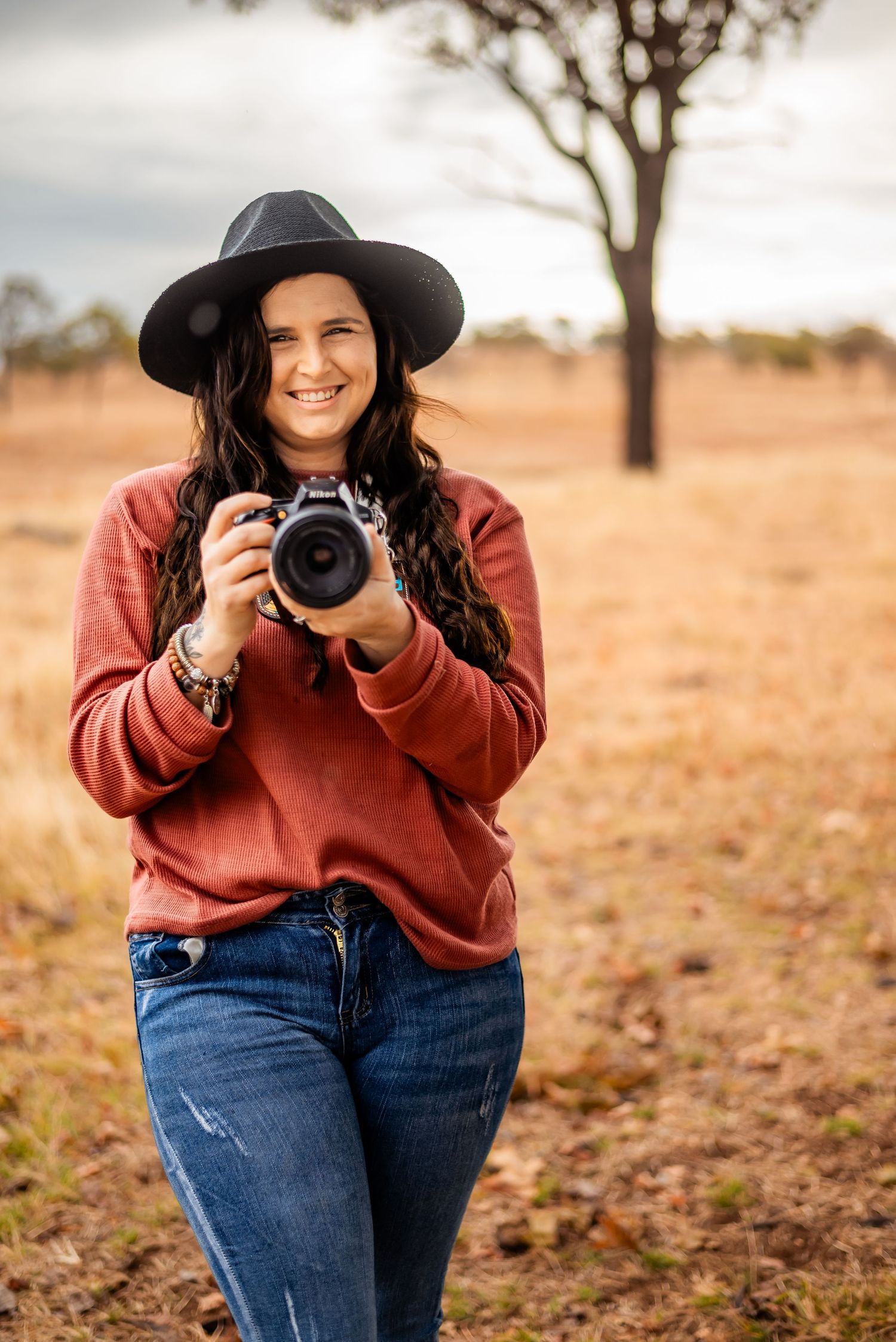 Photographer in hat and sweater holding camera in autumn countryside setting.