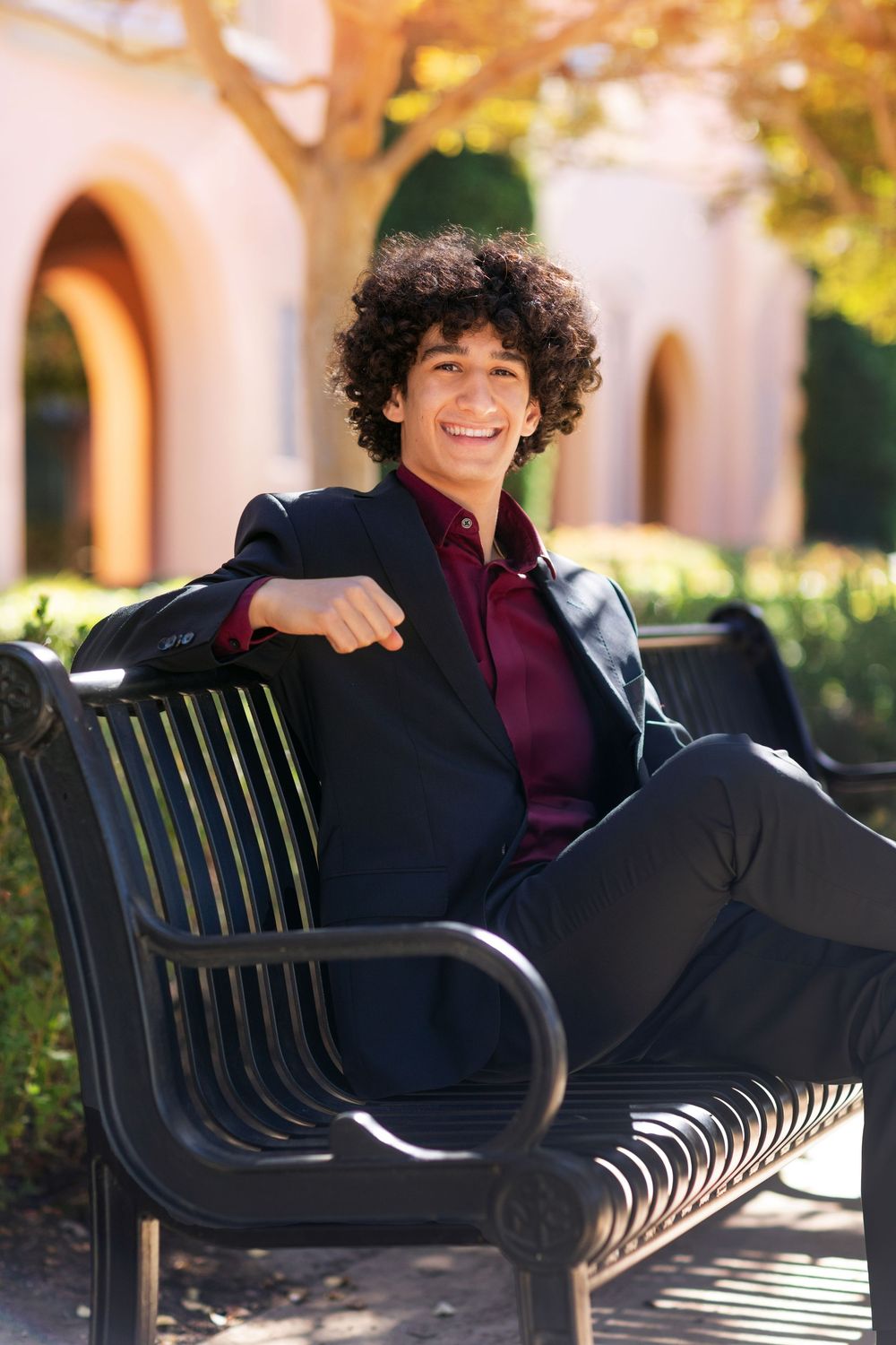 Someone wearing a black blazer and burgundy shirt sits relaxed on a metal park bench on a sunny day.