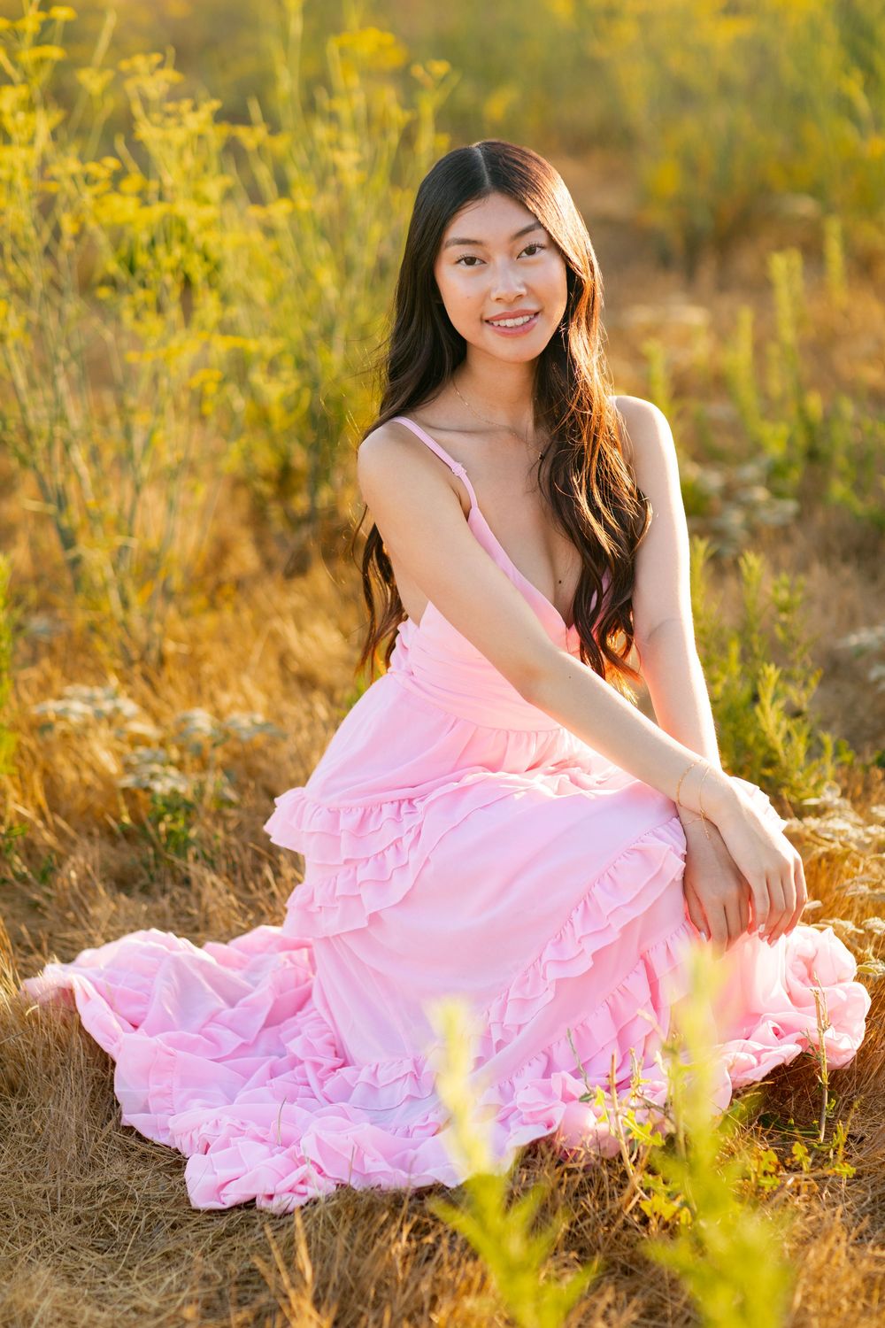 Person in flowing pink dress sits in golden field at sunset surrounded by yellow wildflowers.