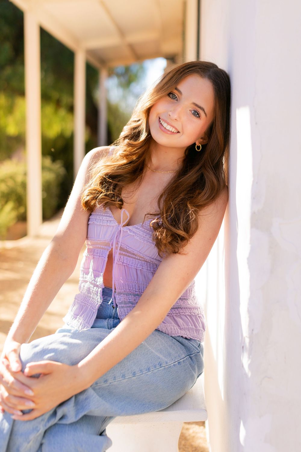 A young woman in a purple crop top and jeans sits on a white porch in warm sunlight.