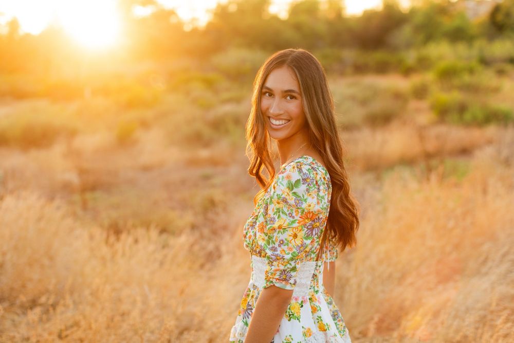 Woman in floral dress stands in golden field at sunset with warm glowing backlight.