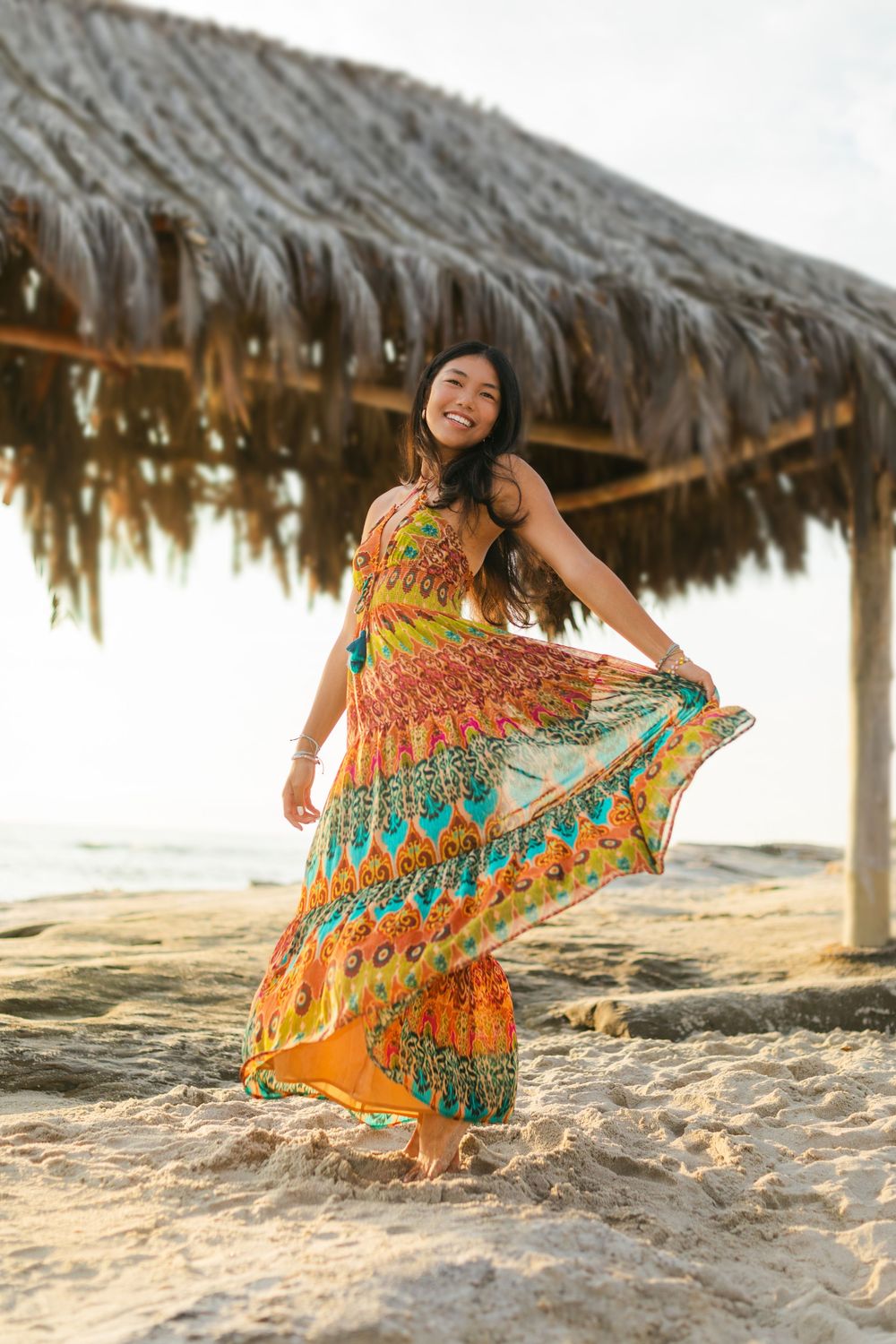 A person in a vibrant orange and yellow floral sundress twirls joyfully on a sandy beach near a thatched umbrella at sunset.