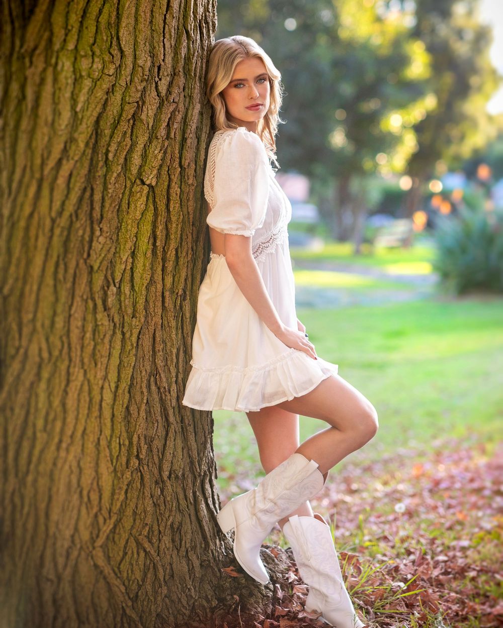 A white lace dress twirls gracefully in a sunlit autumn scene near a large tree trunk with scattered leaves on the ground.