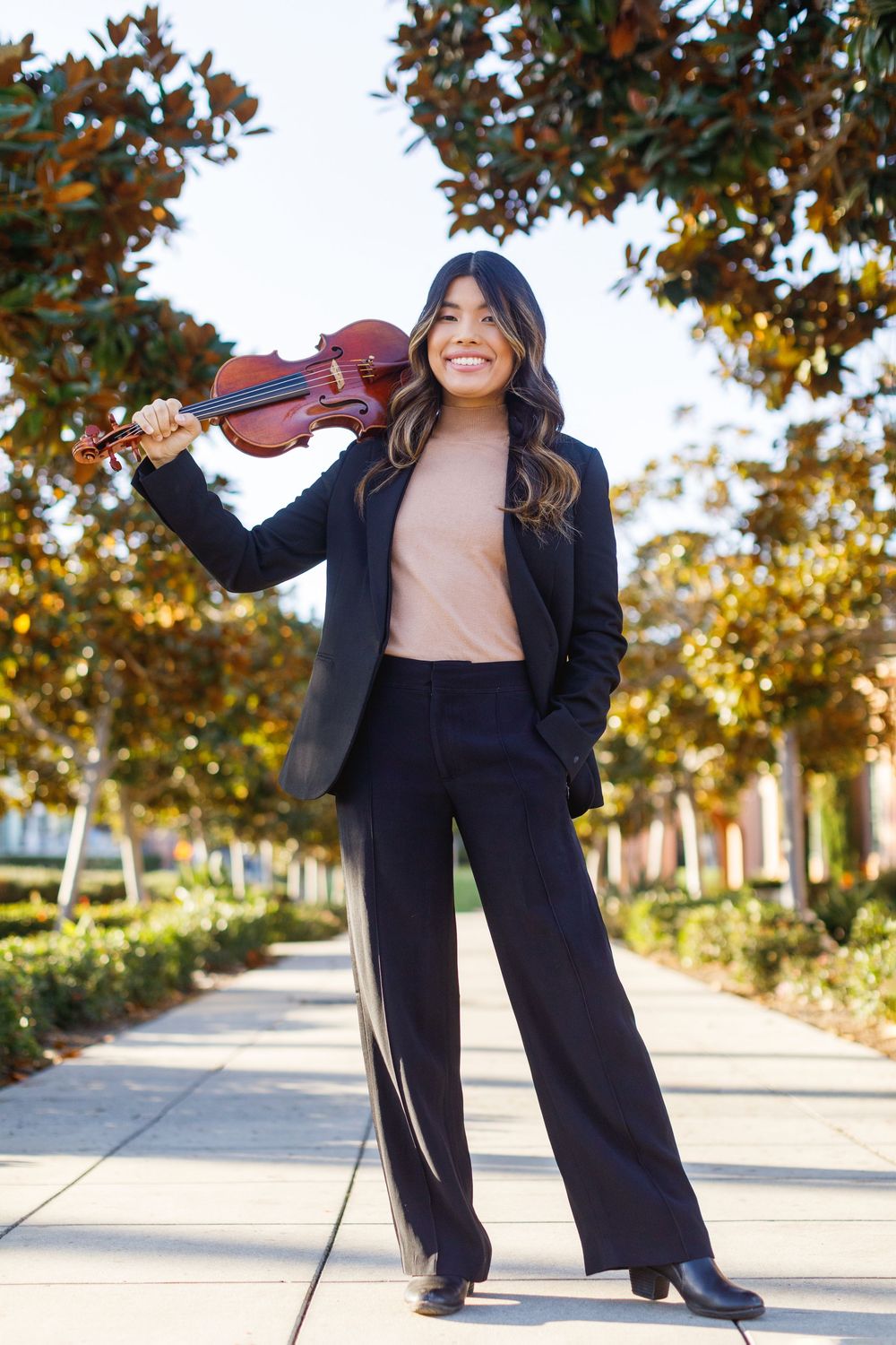 Professional wearing a black blazer and pants strikes playful poses outdoors under autumn trees on a walkway.