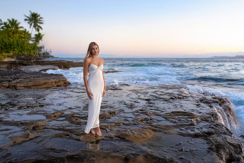 Dreamy sunset photoshoot on a rocky coastal beach with crashing waves and palm trees in the background.