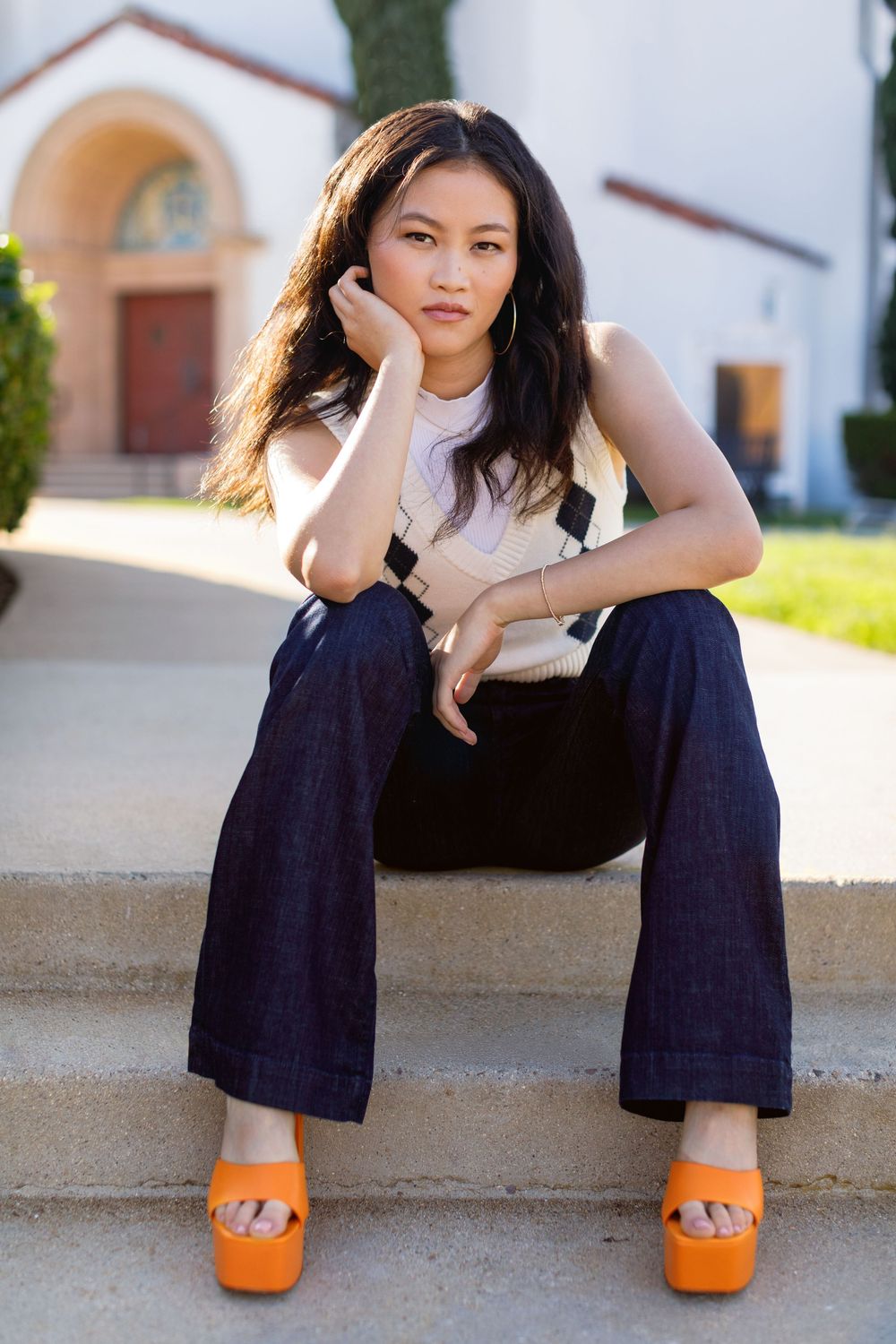 Person in white sleeveless top and dark wide-leg pants sits on steps wearing bright orange platform sandals in sunlight.
