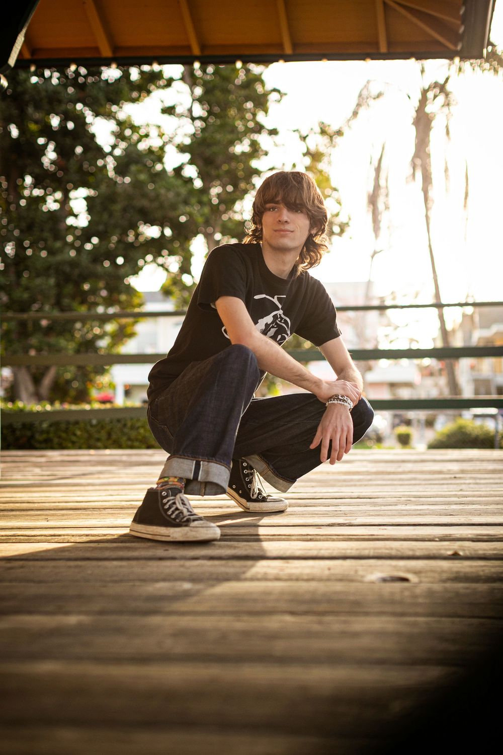 Person in black clothing crouches on wooden deck during golden hour with trees in background.