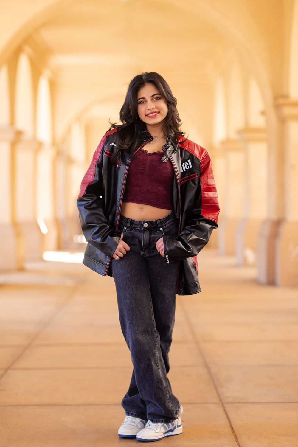 Young person in burgundy crop top and oversized leather jacket poses in arched hallway with warm lighting.