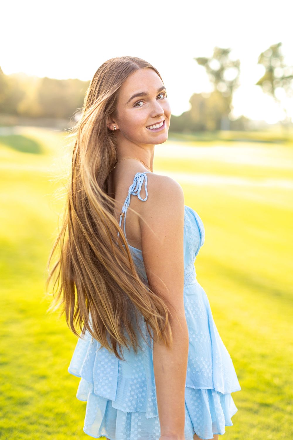 Person in a light blue summer dress smiles joyfully in a sunlit meadow at golden hour.