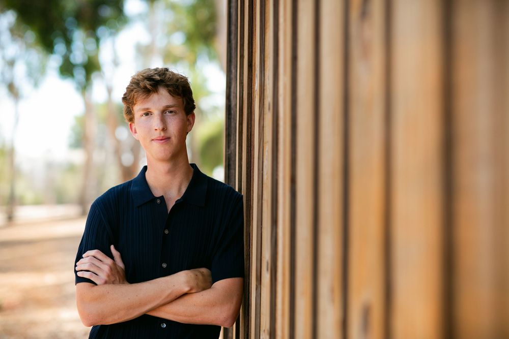A person in a black polo shirt stands with crossed arms against a wooden column in soft outdoor lighting.