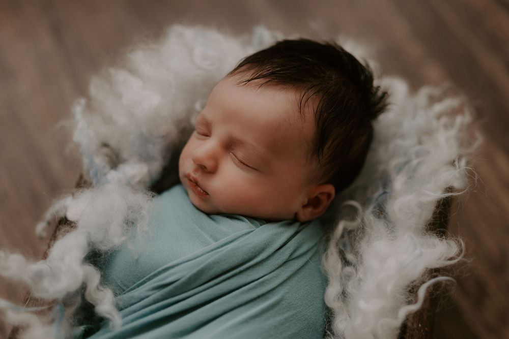 Newborn baby sleeping peacefully wrapped in teal fabric with cloud-like backdrop.