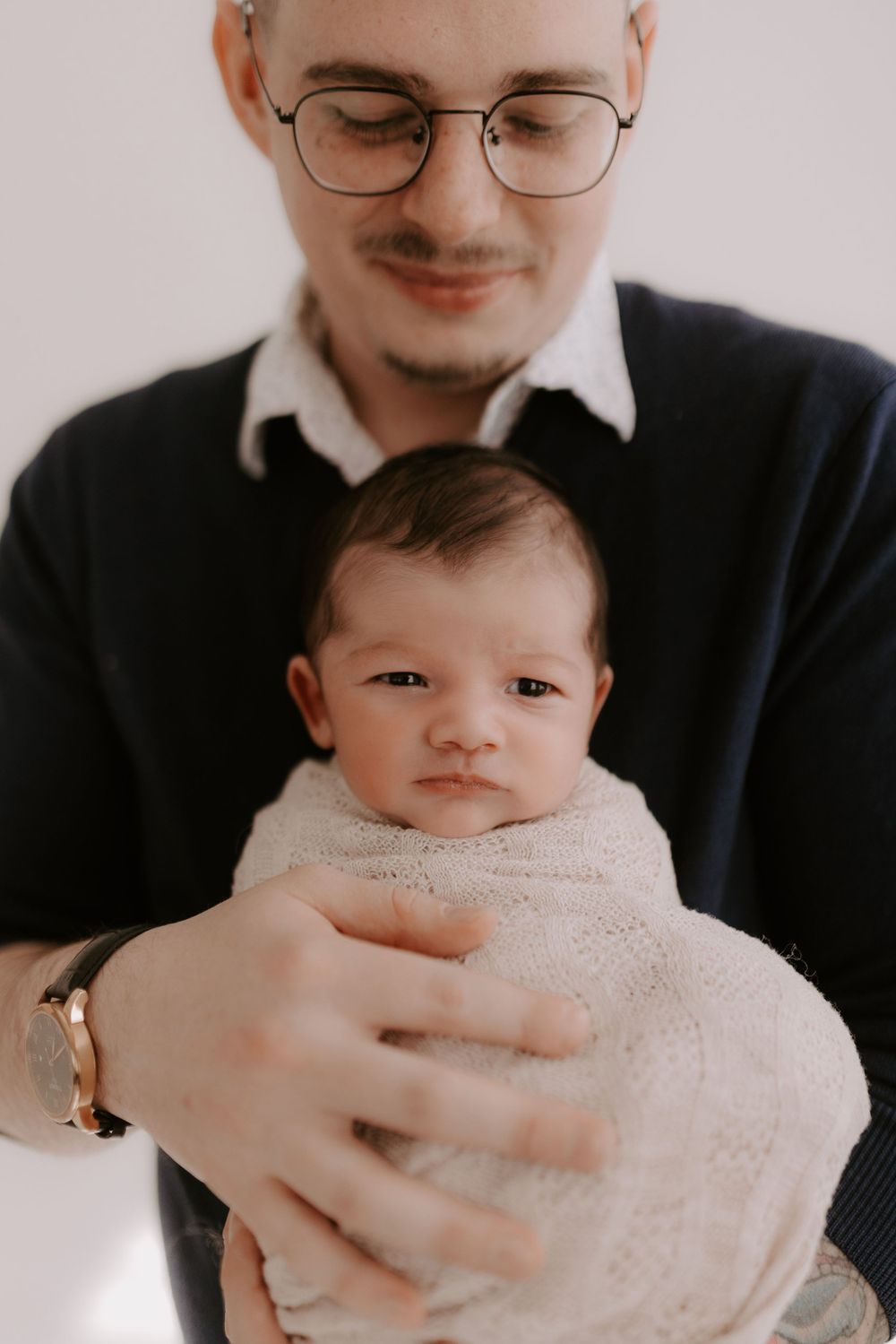 A newborn baby wrapped in a white blanket is held gently in close-up shots.
