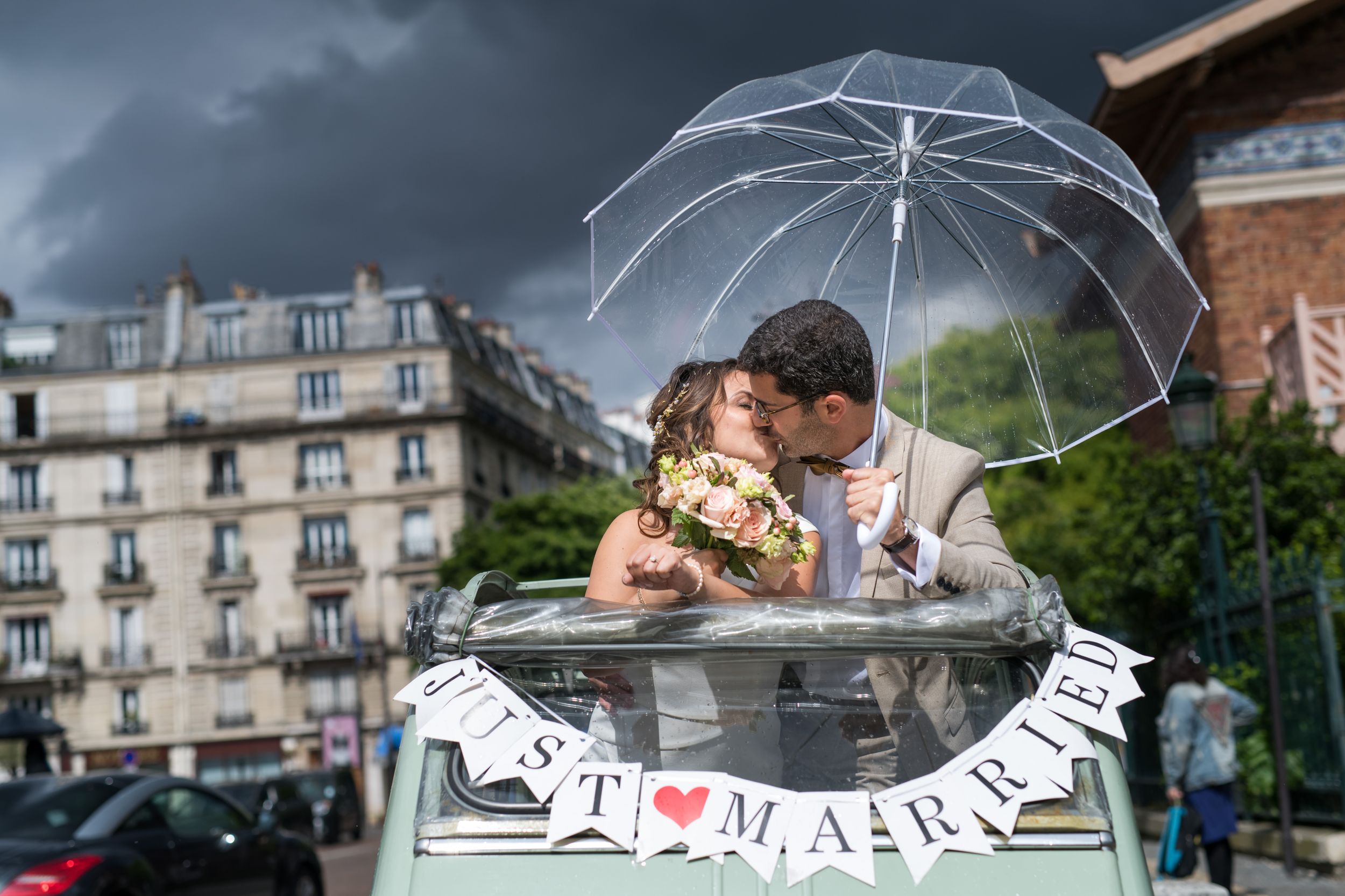 Jeune couple de mariés s’embrassant sous un parapluie transparent par temps de pluie, scène romantique.