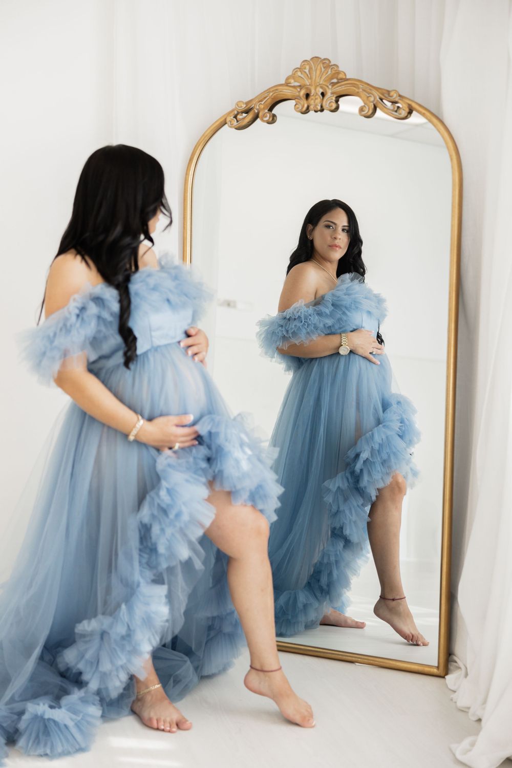 A maternity photo shoot featuring a flowing blue tulle dress and ornate gold mirror against a white backdrop.