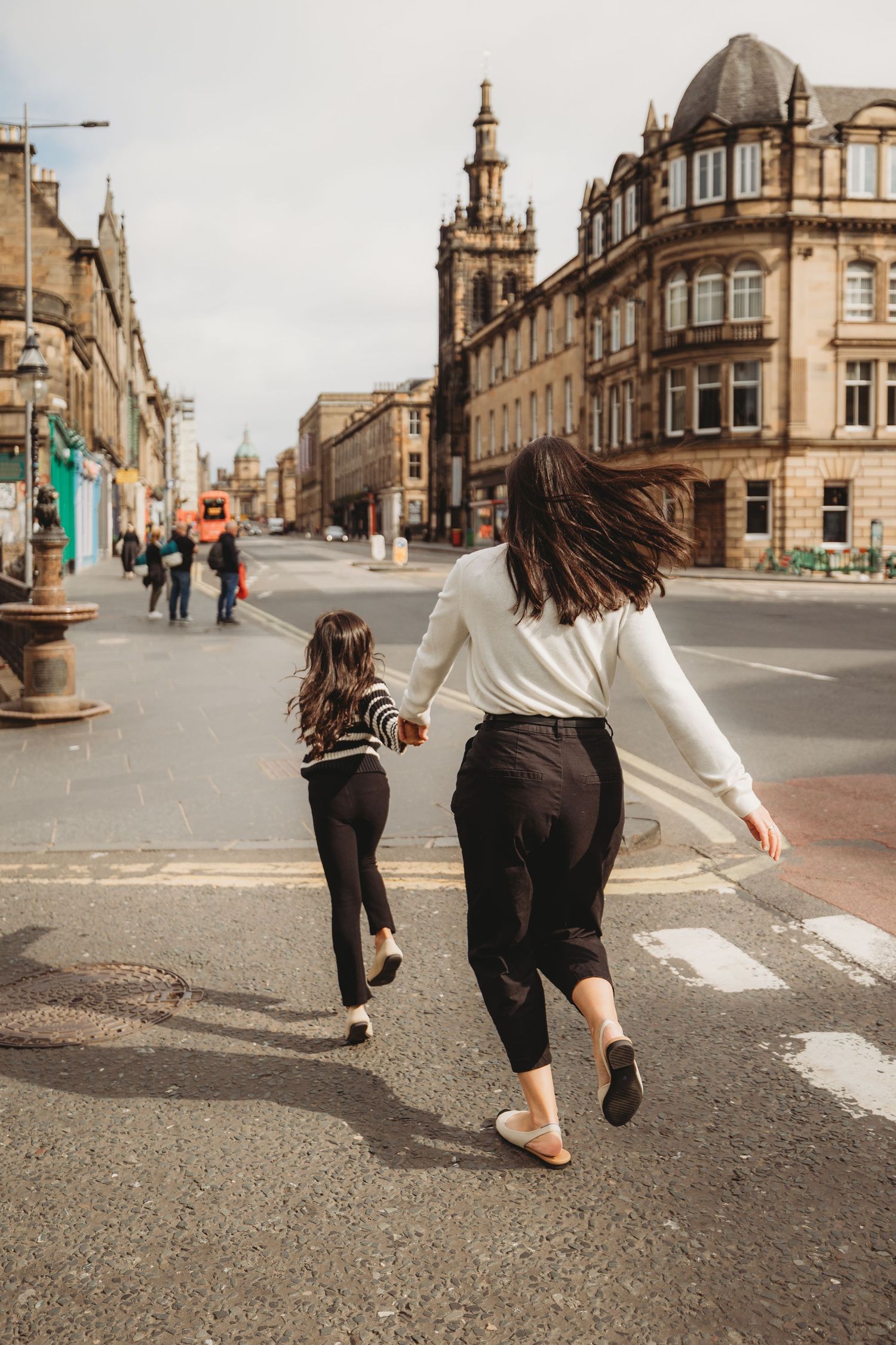 Two people playfully running and dancing through historic city streets with classic stone architecture in the background.