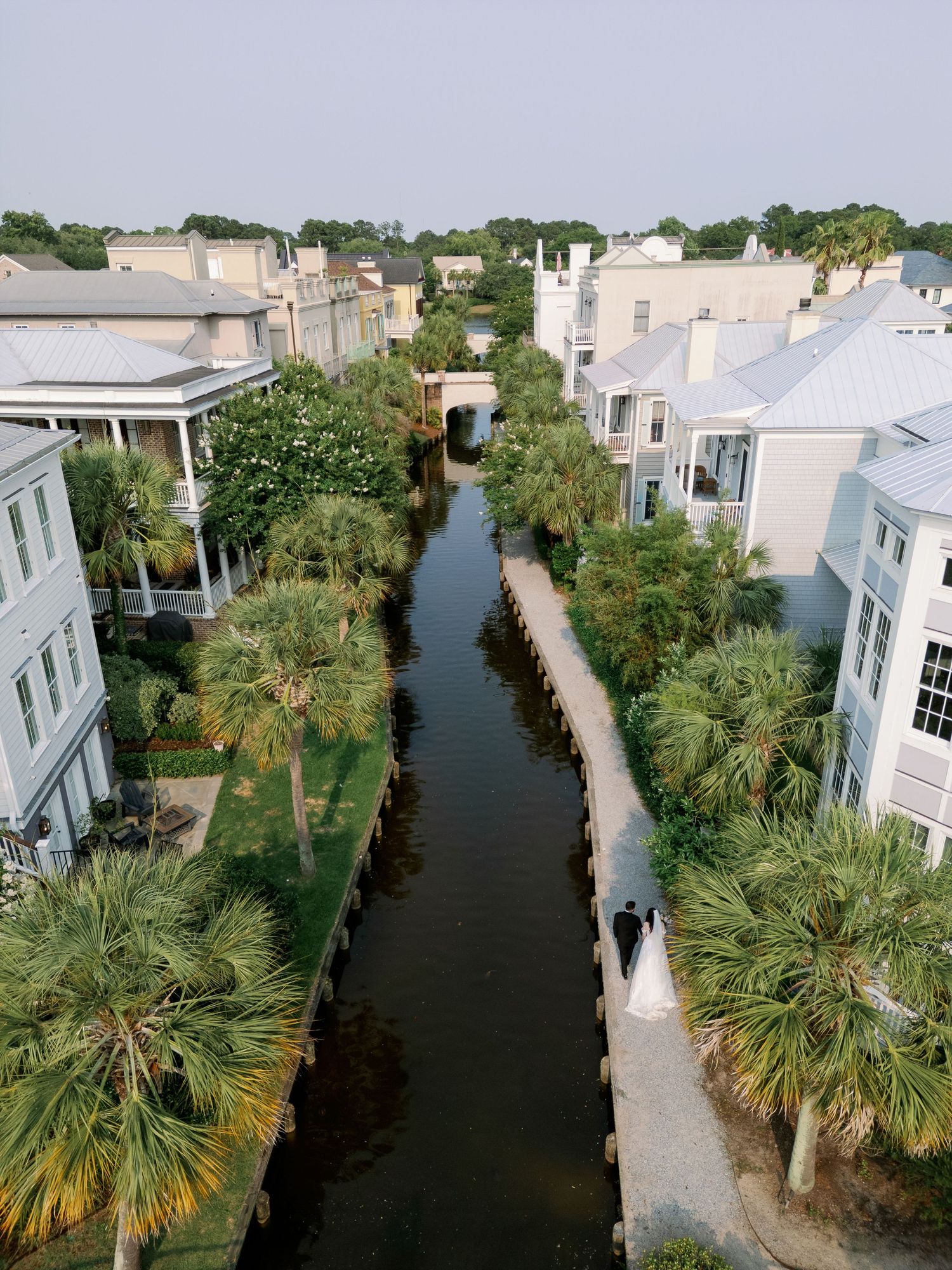 Aerial drone view of bride and groom walking along canal lined with palm trees and pastel homes.