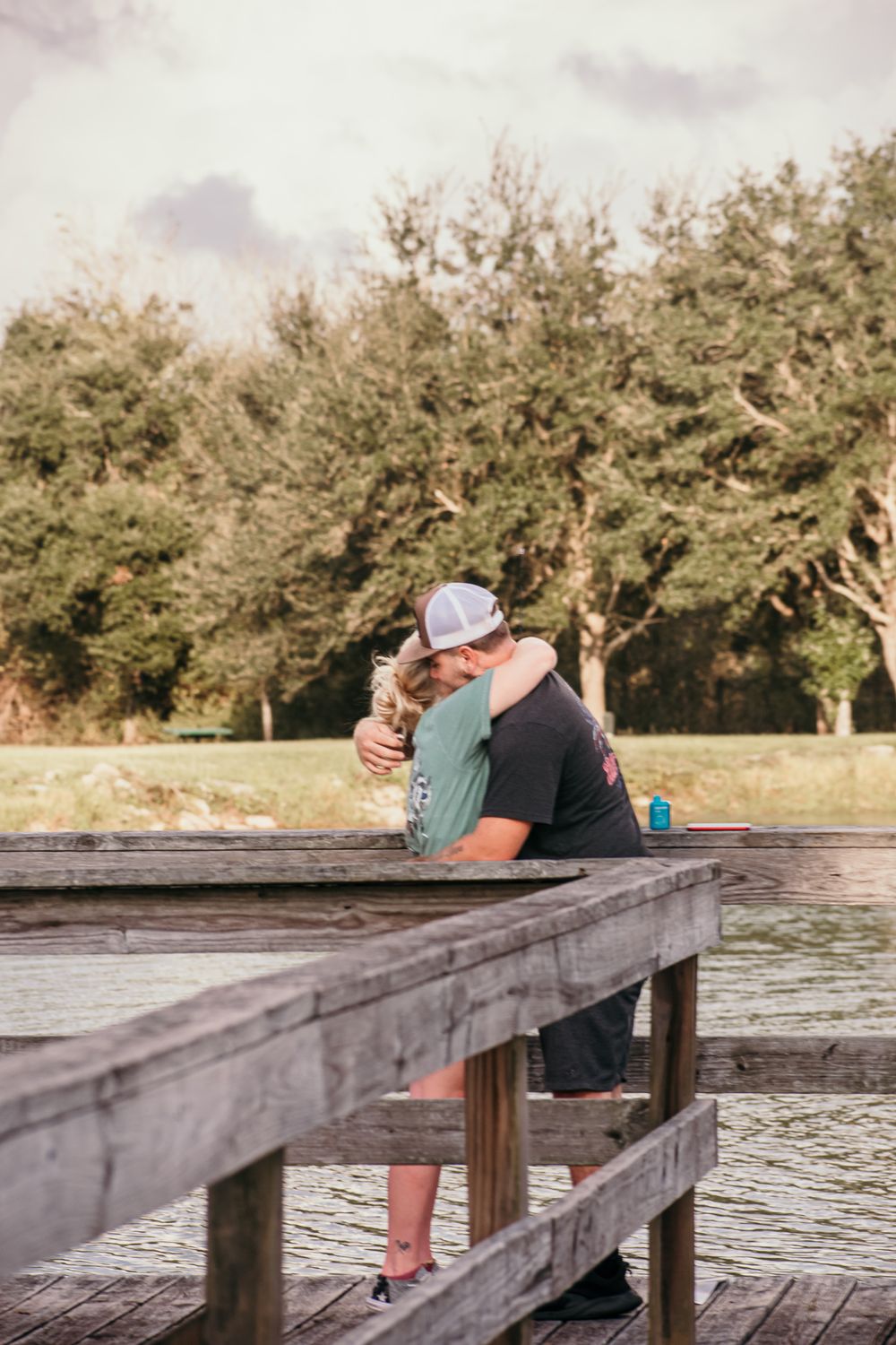 A couple sits together on a wooden dock overlooking a lake during sunset with trees in the background.