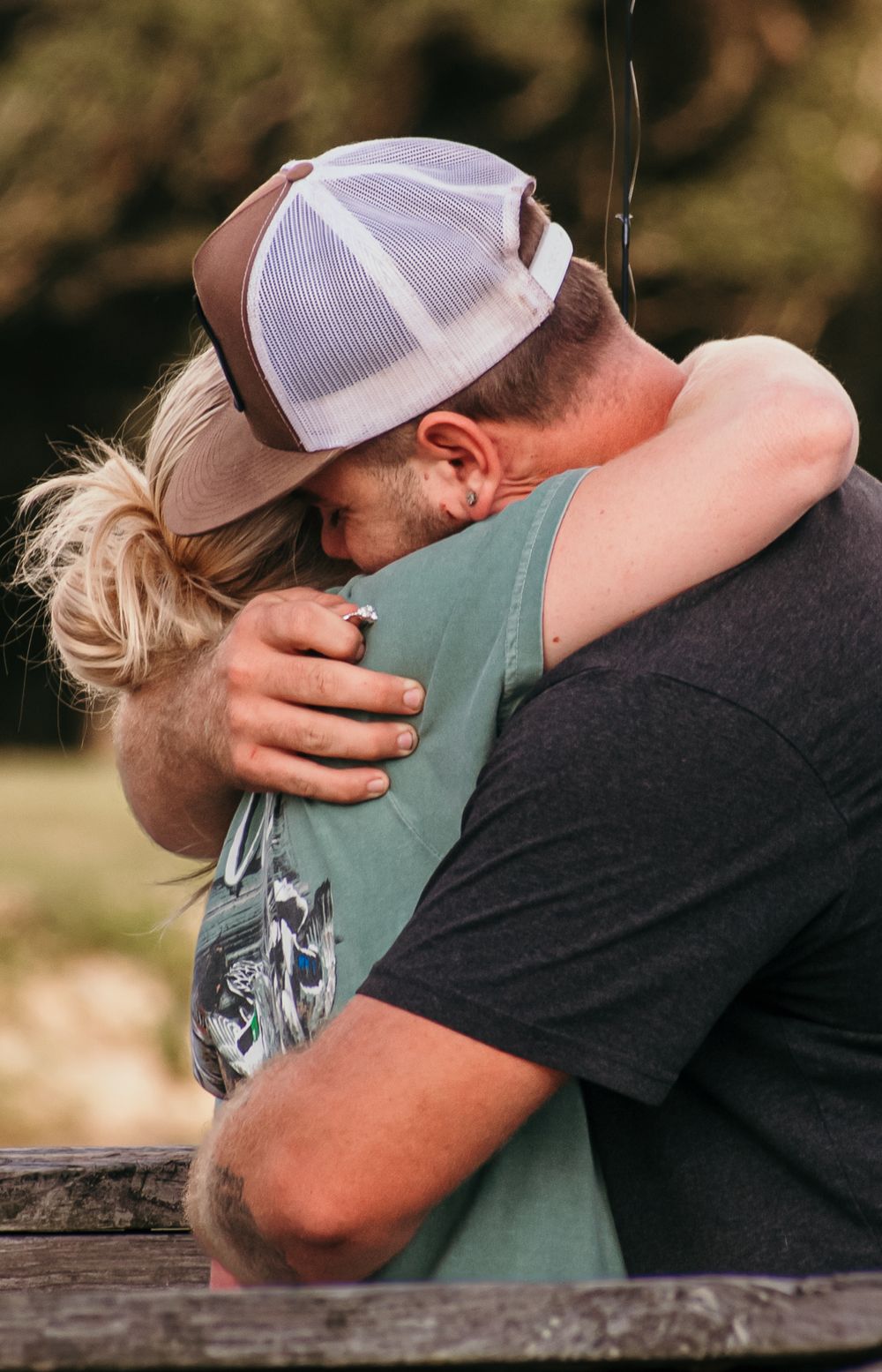 Two people share an emotional embrace outdoors, with one wearing a trucker cap and the other in a sage green shirt.