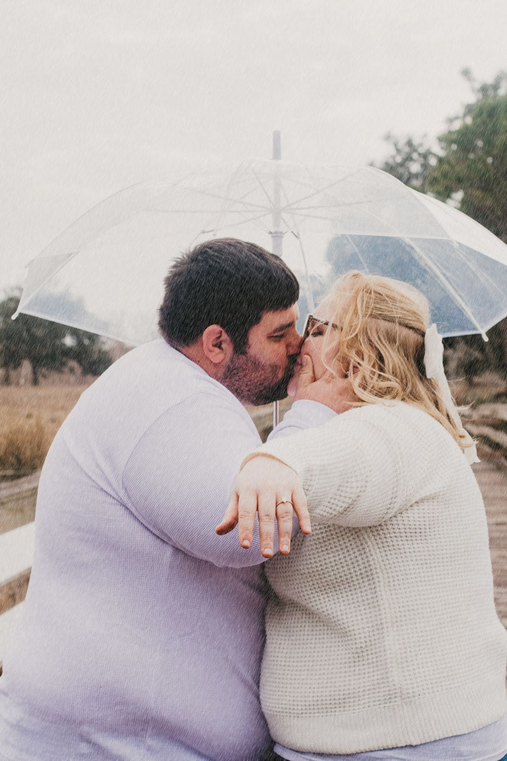 A romantic couple shares a kiss under a clear umbrella in a misty outdoor setting while wearing cozy sweaters.
