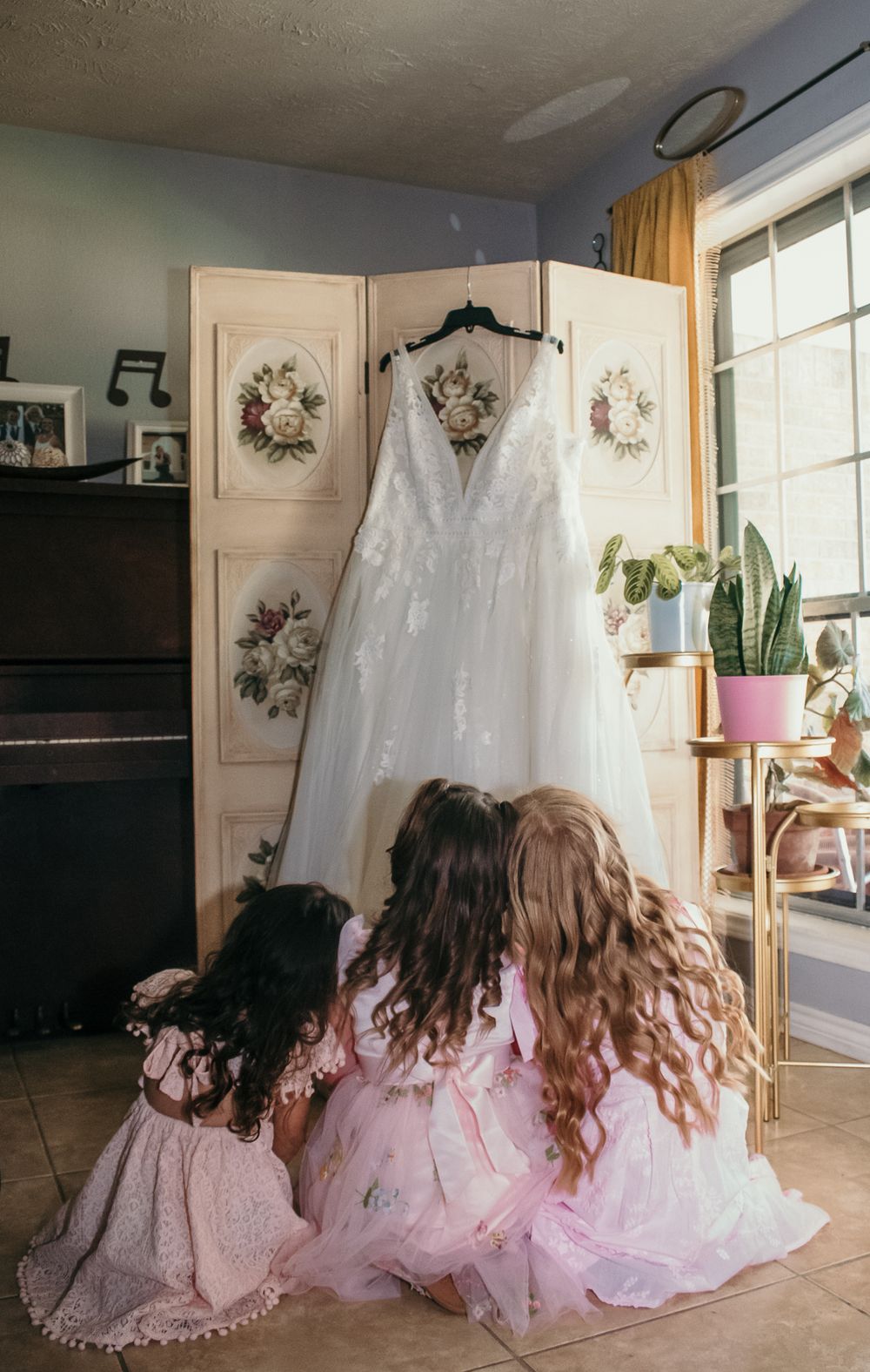 White wedding dress hanging on display with flower-adorned room divider while two people in pink dresses sit on floor.