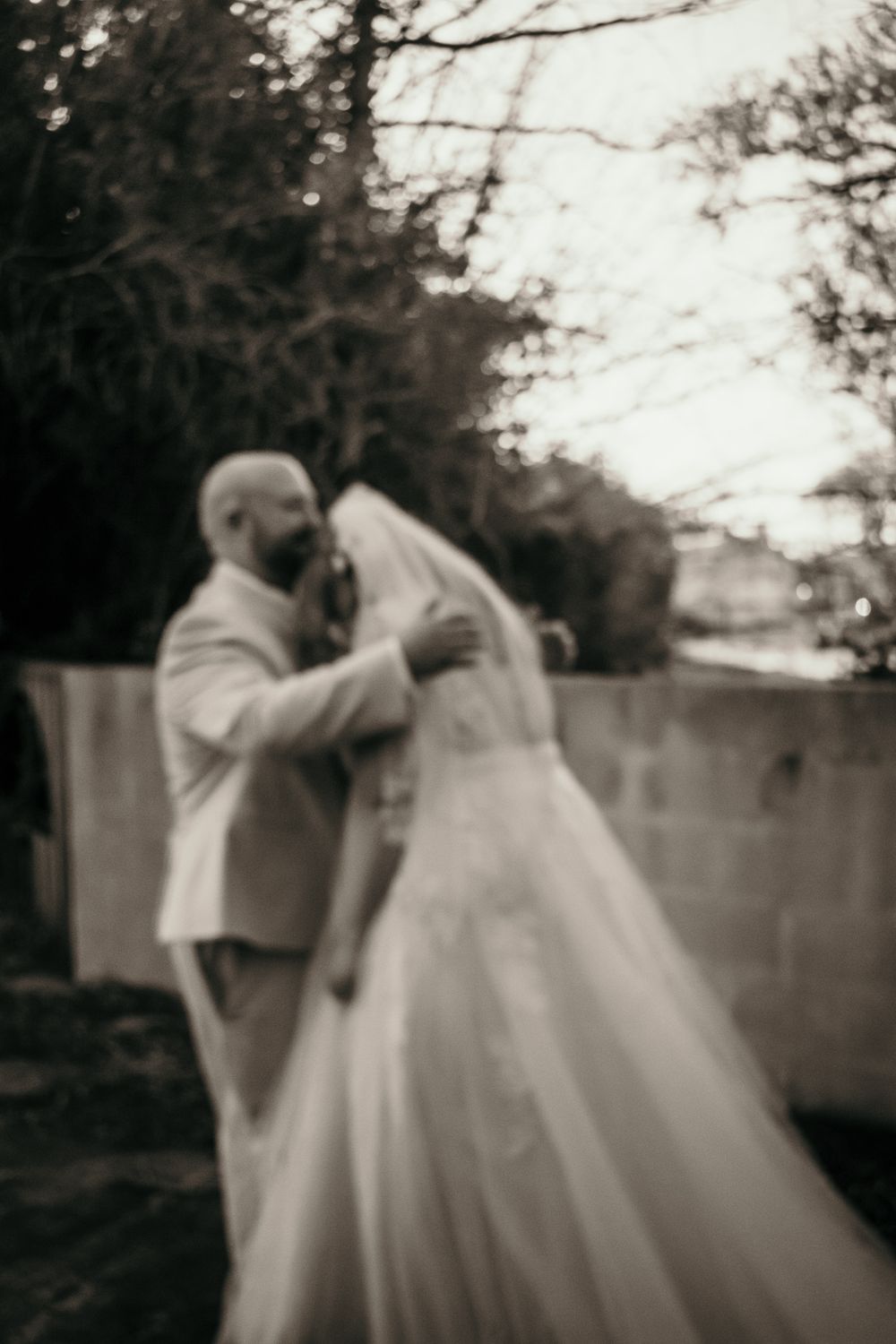 A soft-focus black and white wedding photo showing a romantic moment outdoors with trees in the background.