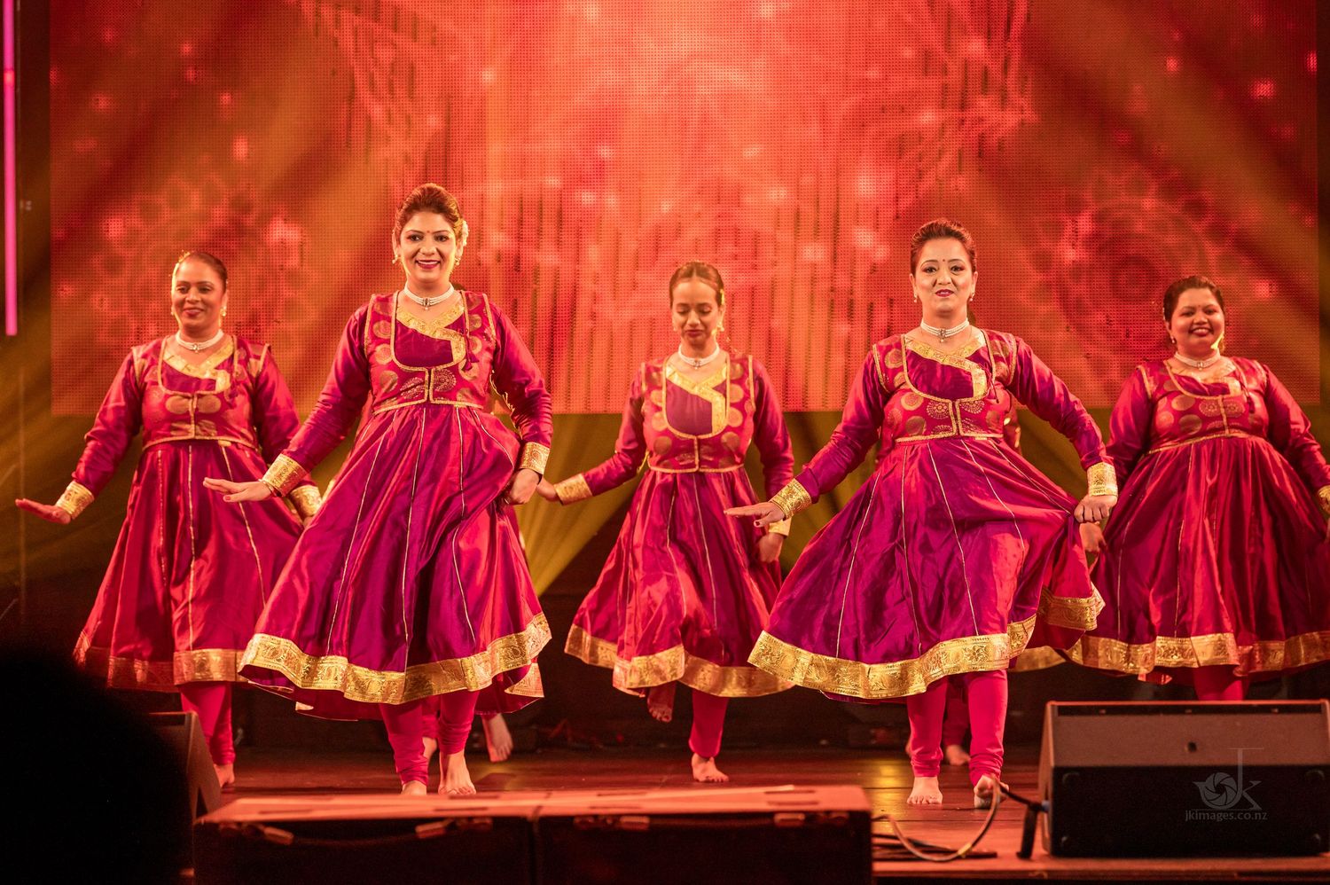 Performers in matching burgundy and gold traditional Indian dance costumes perform synchronized steps on stage with red lighting.