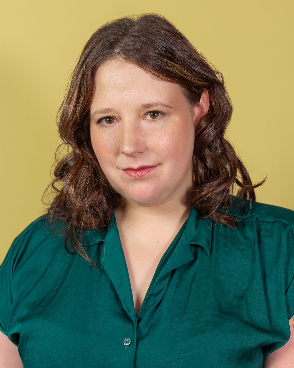 Headshot photography: a woman wearing a green top sits against a yellow backdrop with a serious expression