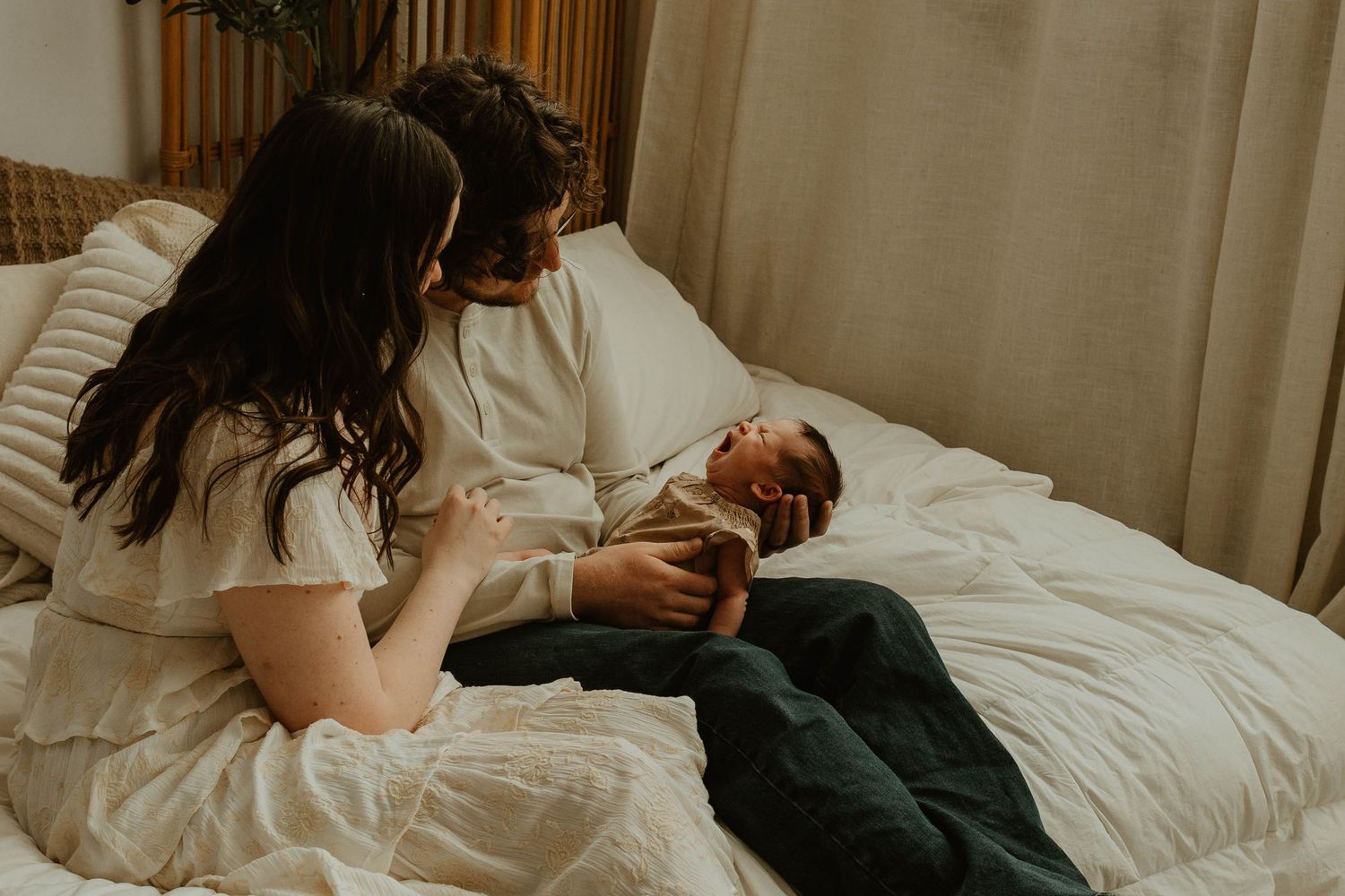 Two people on a bed look lovingly at newborn baby swaddled in blanket while resting on white bedding.