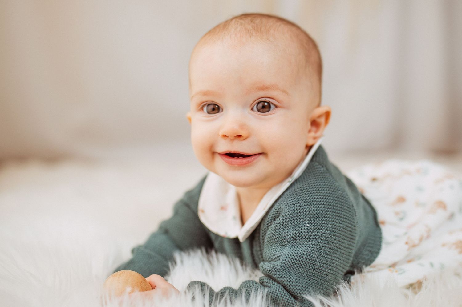 Smiling baby in grey sweater lying on patterned bedding.