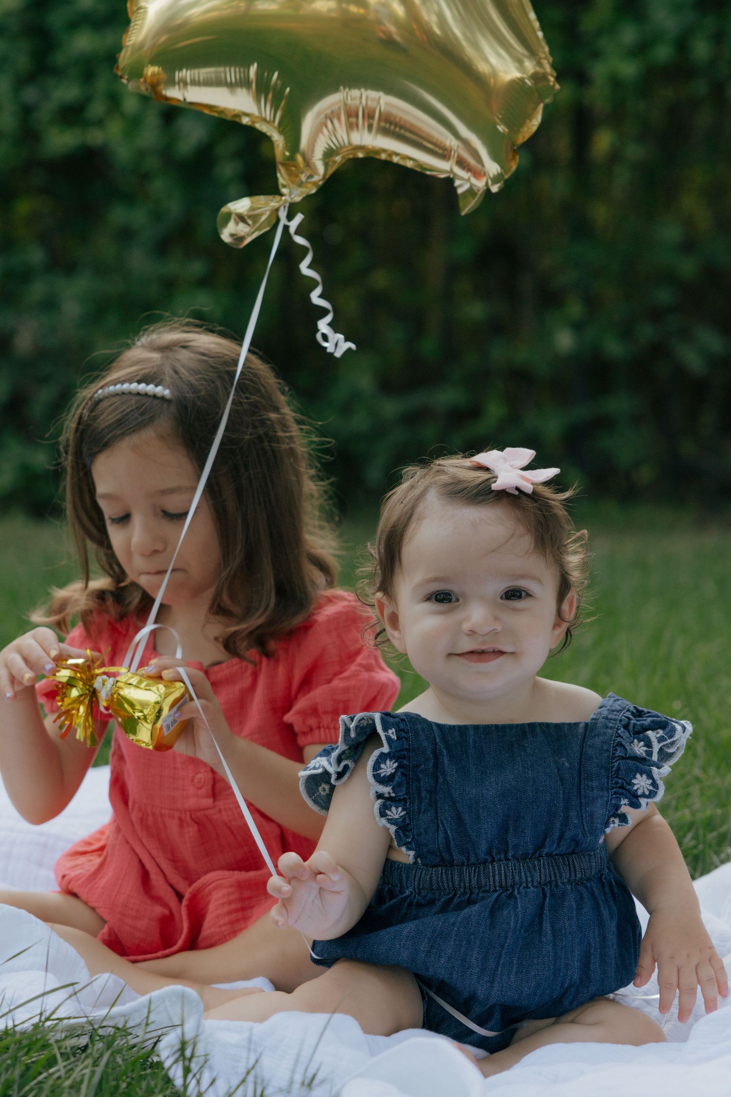 Two children sitting on a white blanket outdoors with a gold balloon on a summer day.